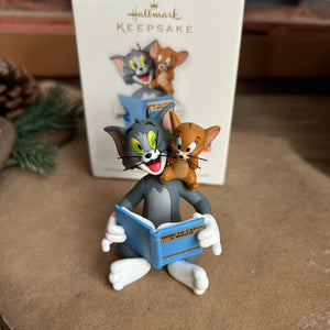 A Hallmark Keepsake ornament featuring cartoon characters Tom and Jerry. Tom is seated and reading a book to Jerry, who is sitting on Tom's lap.
