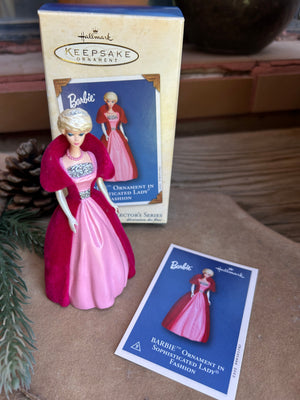 Barbie doll ornament with pink dress and red cape on a surface with packaging and collectors card 