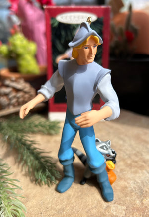A collectible keepsake ornament of Captain John Smith from the Pocahontas Disney movie, wearing a blue outfit and standing next to meeko the sweet raccoon.
